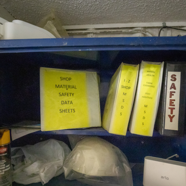 A picture of a metal blue cabinet containing several binders, one displayed that says "SHOP MATERIAL SAFETY DATA SHEETS" on the cover. 