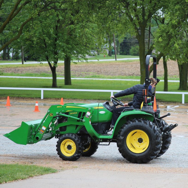 A man looks backward as he backs up a green tractor with bucket loader attached in a gravel lot.