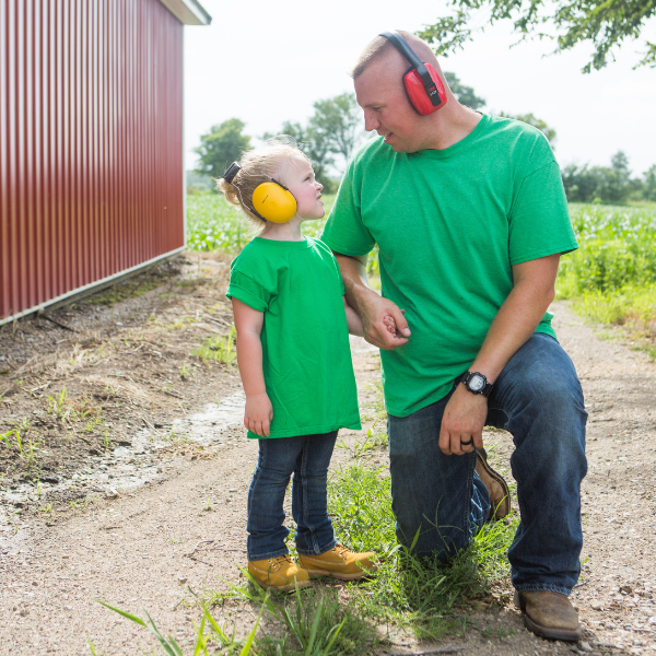 A man kneeling by a little girl, both are in green t-shirts, jeans, boots, and are wearing hearing protection.