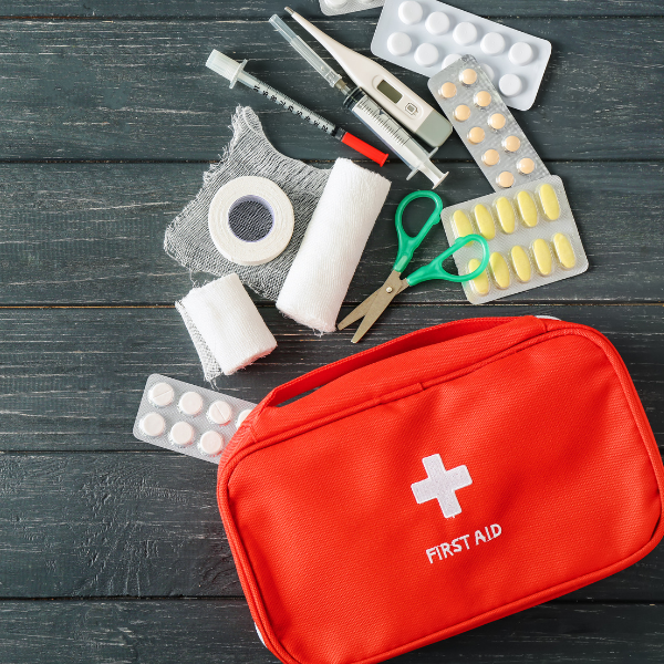 A red first aid kit is unzipped with materials spilling out, including gauze, scissors, a syringe, thermometer, and blister pack of medication.