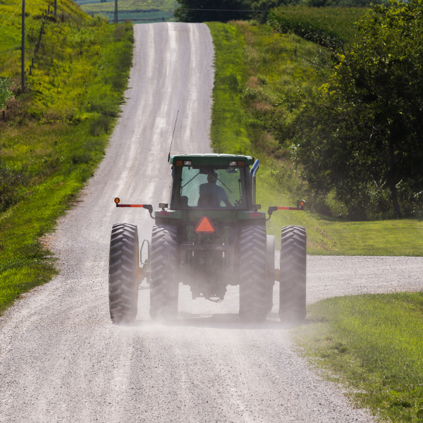 A green tractor is driving away from the camera on a gravel road, with dust surrounding it and a slow moving vehicle sign on the back. 