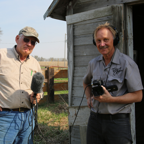 Two men stand outside a weathered barn, one holds a sound boom and one holds a camera while wearing headphones.