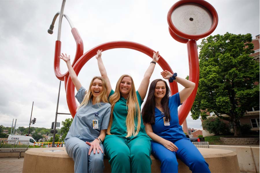 Three female students in scrubs pose in front of a sculpture of a stethoscope