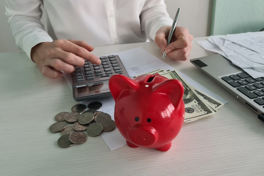 person using a calculator, coins, money bills, piggy bank, laptop, papers