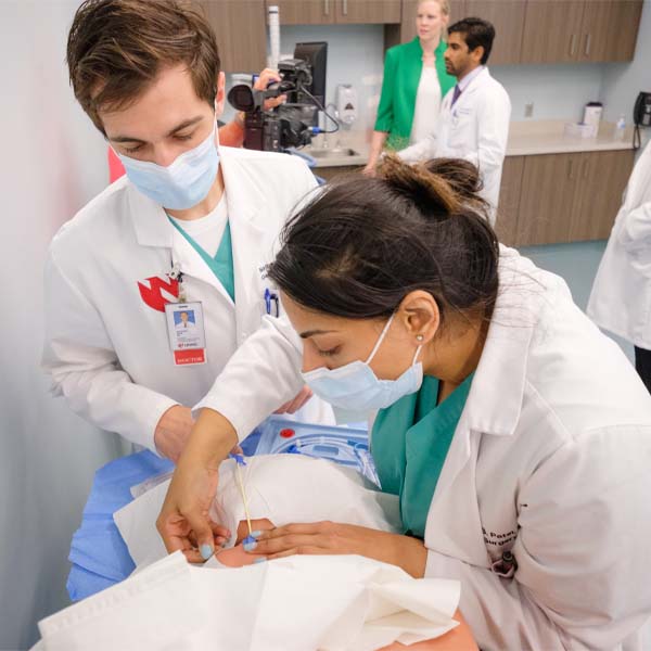 Two students in white coats work on a mannequin