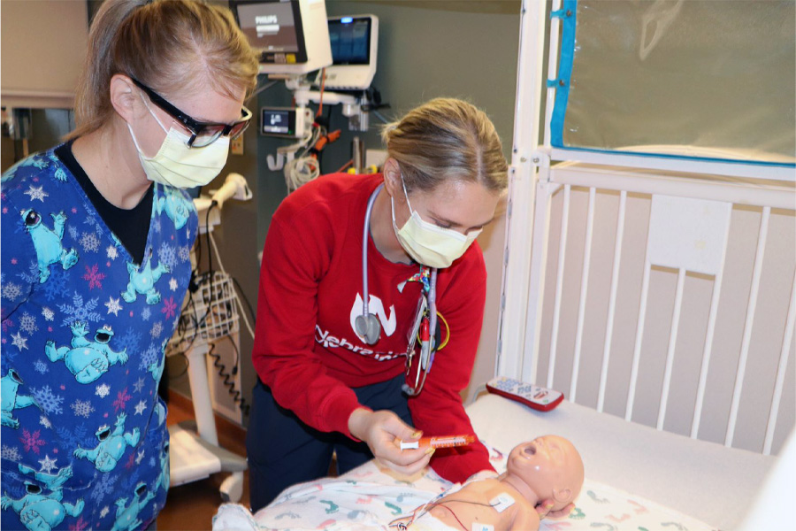 A female health care worker watches as a female job shadow student works with an infant mannikin