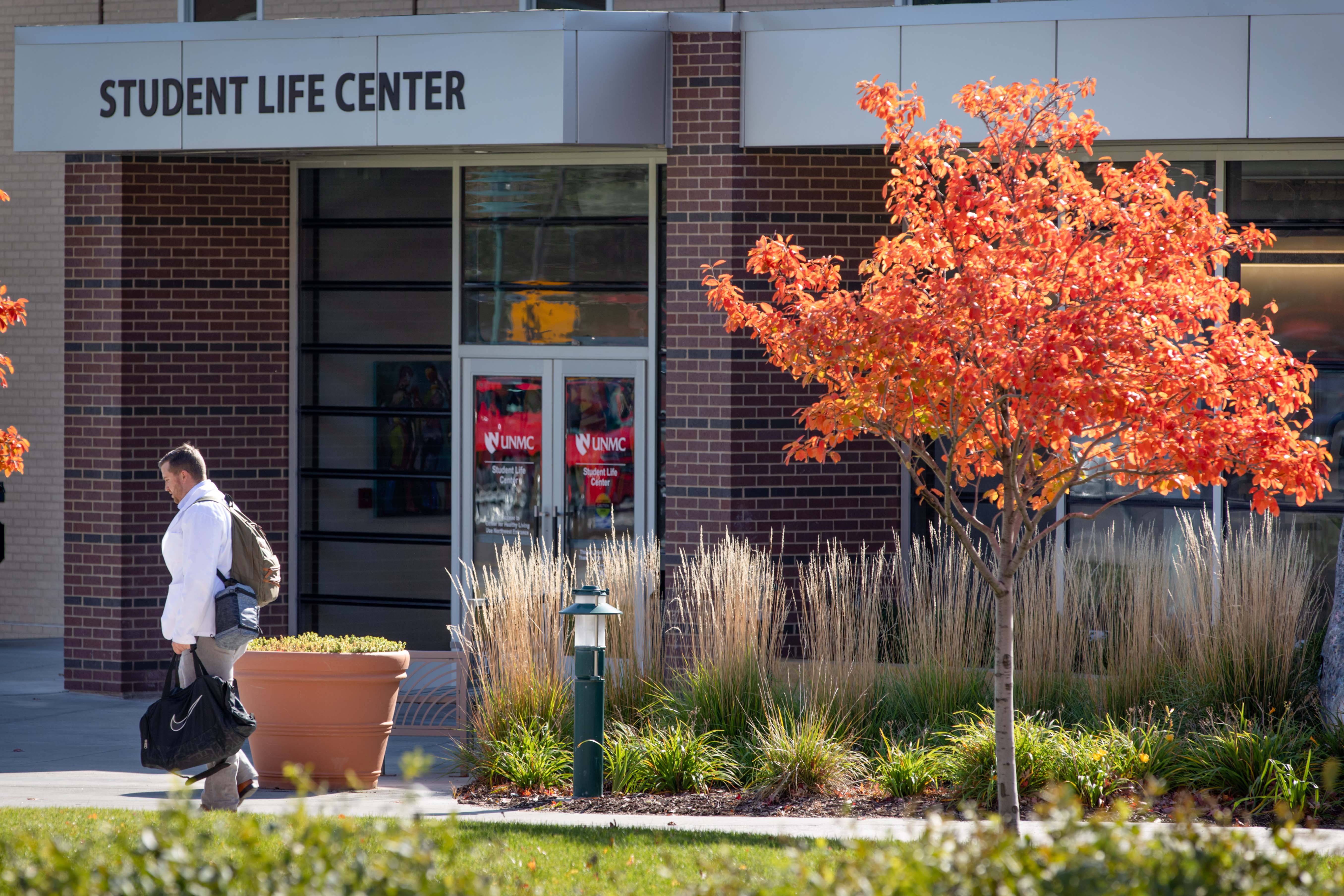 a male student walks in front of the Student Life Center, fall tree with orange leaves