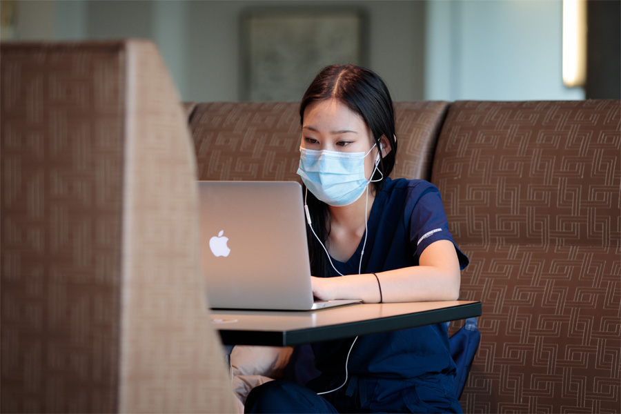 A student wearing scrubs and a mask works at a laptop