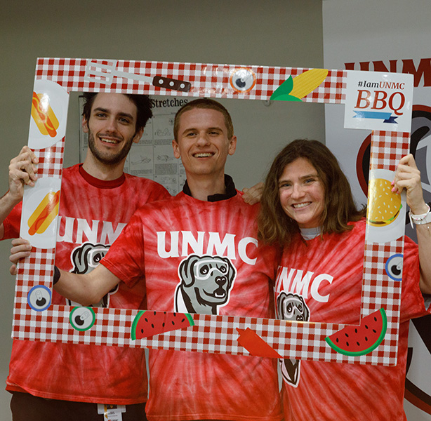 three students wearing UNMC T-shirts with Ellie the Lab on the front, posing inside a cardboard picture frame at the barbeque