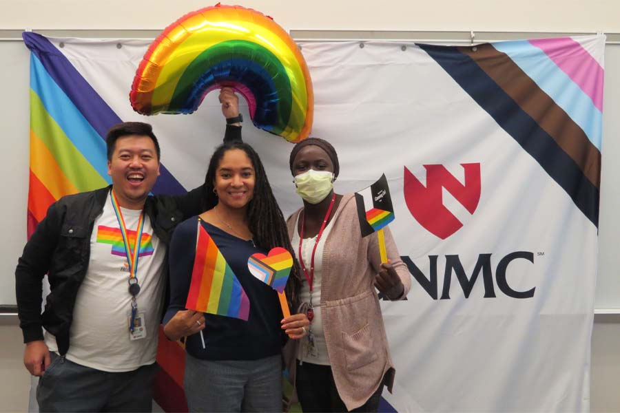 Three people pose in front of a rainbow background with the UNMC logo