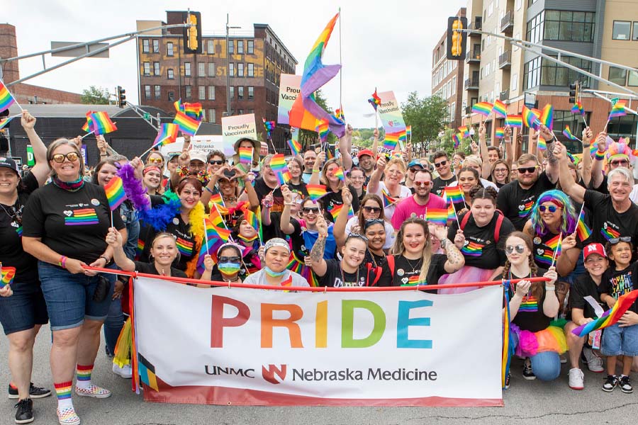 A large group waves rainbow flags in front of a banner reading "Pride, UNMC, Nebraska Medicine"