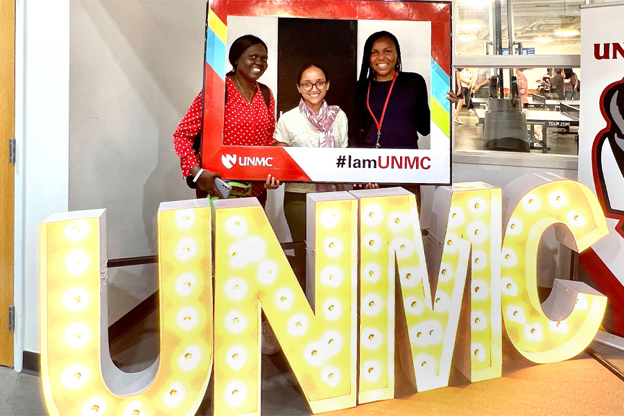 Three students stand behind lighted letters spelling out "UNMC" while holding a cardboard frame with the UNMC logo and text "#IamUNMC"