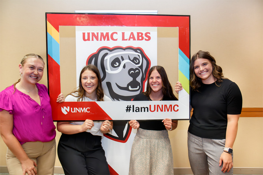 Four students pose with a cardboard frame with the UNMC logo and text "#IamUNMC"