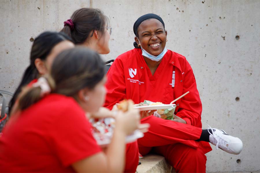 Four UNMC nursing students in red scrubs eat food from the UNMC BBQ