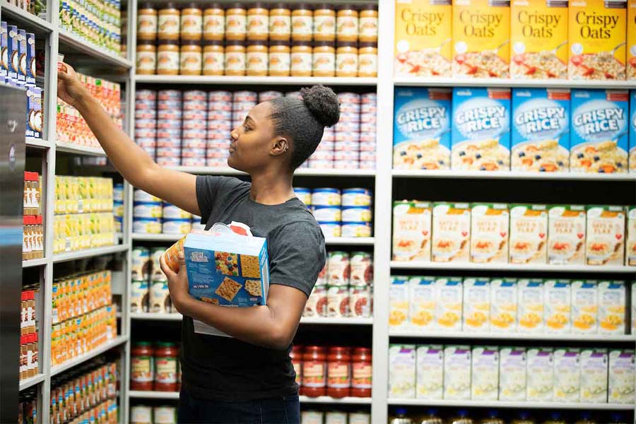 A woman in a food pantry holding boxed food items reaches for an item on a shelf