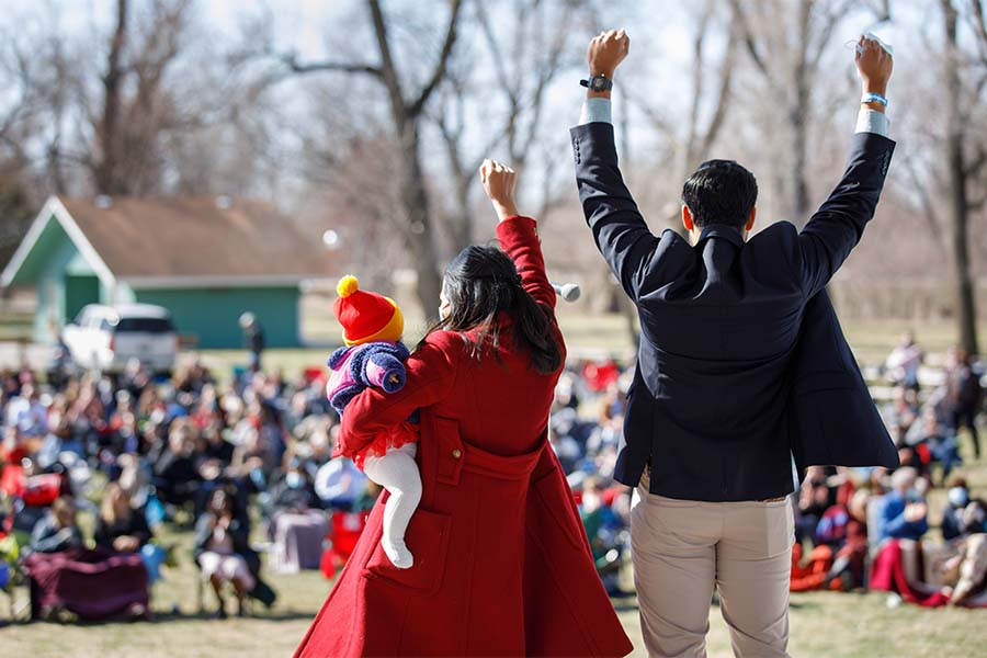 A woman holding a baby and a man hold their hands up in celebration in front of a crowd