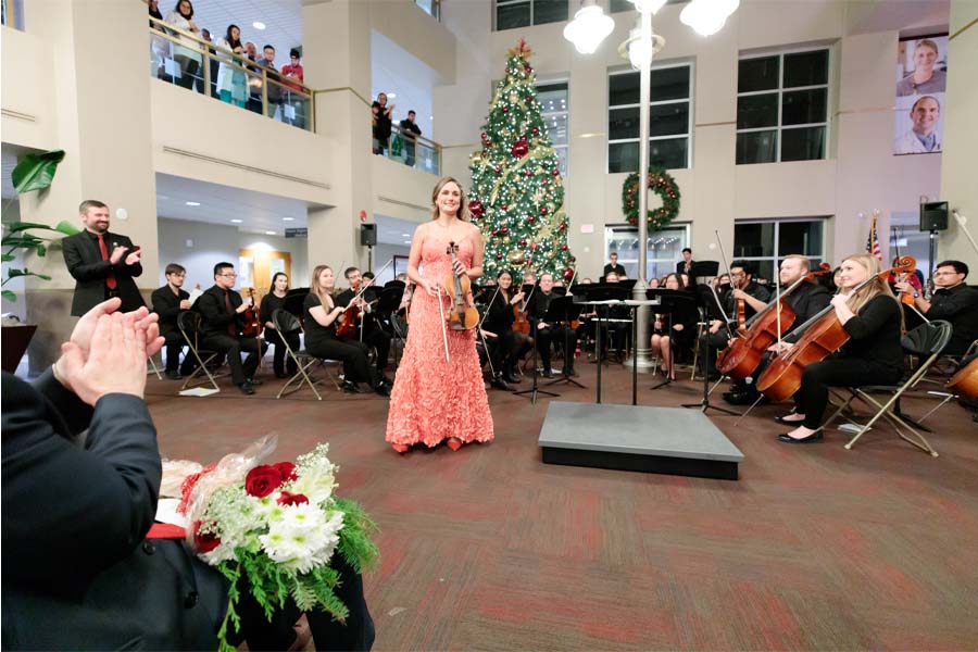 A woman in a pink dress holds a violin in front of a crowd, with a large Christmas tree in the background