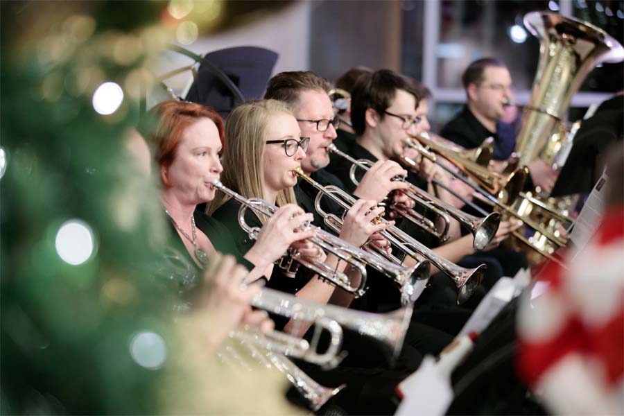 A group of people play trumpets and a trombone, with a Christmas tree in the foreground